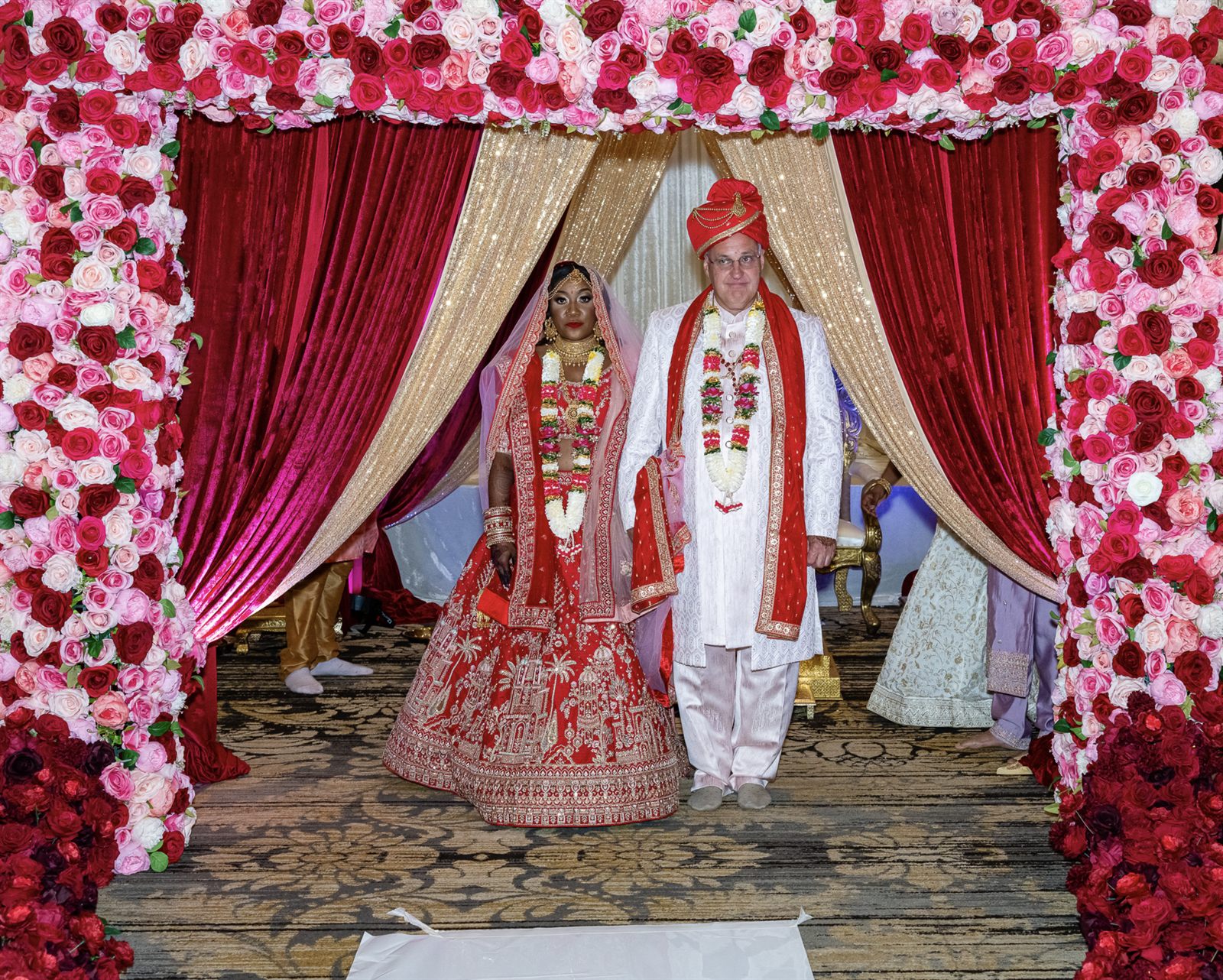 A couple in traditional attire under a rose floral arch